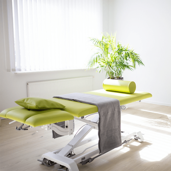 Eine helle Physiotherapiepraxis mit einem grünen Behandlungsbett, einem grauen Handtuch und einer Pflanze.