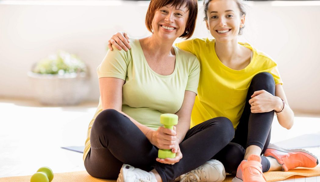 Zwei Frauen in gelber Sportkleidung sitzen lächelnd auf einer Yogamatte, eine hält eine grüne Hantel.