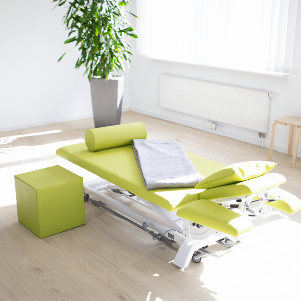 Eine grüne Therapieliege steht in einem hellen Raum neben einem großen Pflanzenkübel.