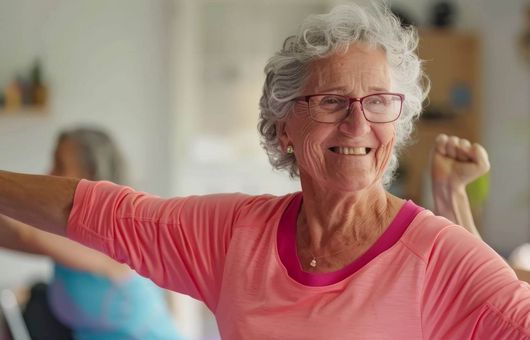 Eine glückliche Seniorin in einem rosa Oberteil genießt lächelnd eine Fitnessstunde mit anderen älteren Menschen.