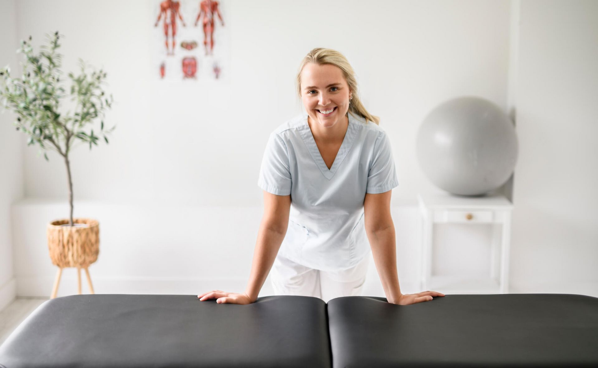Eine lächelnde Physiotherapeutin steht an einem Behandlungstisch in einem hellen, modernen Raum mit einem Gymnastikball.